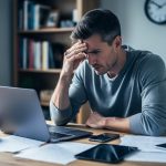 Freelance writer rubbing temples at a cluttered home desk with laptop, smartphone, and tablet; soft daylight from a side window; blurred bookshelves, wall clock, and calendar in the background with no readable text.
