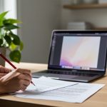 Hands of a freelance editor holding a red pencil over printed pages beside an open laptop on a wood desk, with a leafy plant and reusable mug, in soft natural light and a softly blurred home office background.