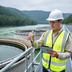 Environmental engineer wearing a hard hat and high-visibility vest holds a water sample and tablet on a catwalk beside circular clarifier tanks at a mine water treatment plant, with a green river and forested hills in the background under bright overcast light.