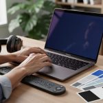 Hands typing on a laptop next to a refreshable Braille display, over-ear headphones, and color swatches on a wooden desk, with a softly blurred home office in the background.